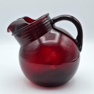 May include: A deep red glass pitcher with a spherical body, a wide pouring spout, and a curved handle. The glass features a translucent finish with horizontal ribbed detailing around the neck area, providing a distinct texture to the vessel.