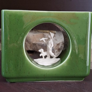 May include: A green ceramic vase with a rectangular shape and a circular opening. Inside the opening is a white figurine of a person with a dog. The figurine depicts a person carrying a tool, with a dog at their feet.
