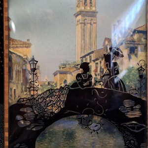 May include: A framed artwork featuring a Venetian scene. Silhouetted figures stand on a bridge with ornate details, set against a backdrop of buildings and a tall tower. The artwork has a brown frame and a vintage aesthetic.