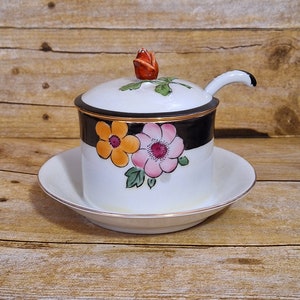 May include: A white ceramic sugar dispenser with a black and gold trim. The dispenser has a lid with a small red rose on top and a matching spoon. The dispenser is decorated with colorful flowers.