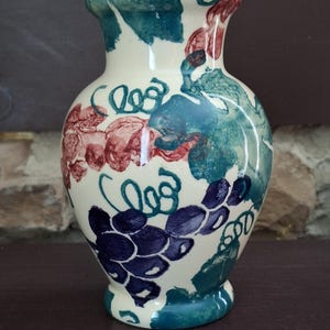 May include: A cream-colored ceramic vase with a painted grape design in shades of blue, red, and green. The vase has a rounded body, a flared rim, and a small base. The grape clusters and vines are hand-painted.