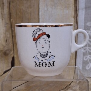 May include: White ceramic mug with a gold rim and a handle. The mug features a cartoon illustration of a woman wearing a hat, with the word "MOM" printed below the illustration. The mug is on a clear glass surface.