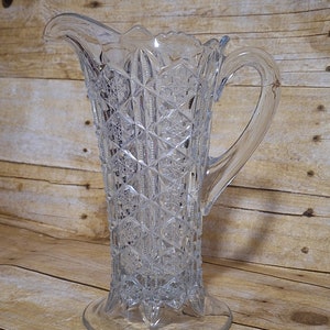 May include: A clear glass pitcher with a cut glass design. The pitcher has a wide base and a tall, slender body. The handle is attached to the side of the pitcher.