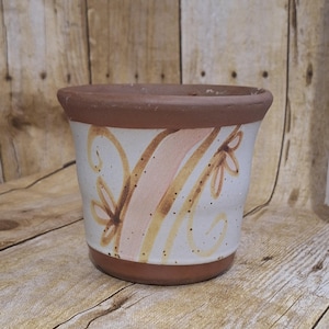 May include: A ceramic flower pot with a white base, adorned with brown, peach, and yellow floral designs. The rim and base are a dark brown color. The pot is sitting on a wooden surface.