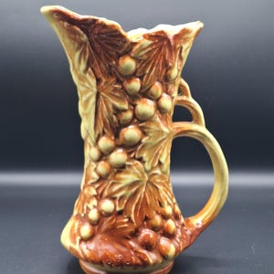 May include: A ceramic pitcher with a handle, featuring a relief design of grapes and leaves. The pitcher is primarily brown and tan, with a flared top and a rounded base. The pitcher is approximately 8 inches tall.