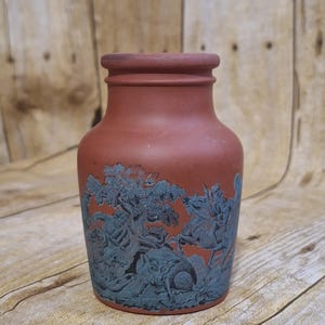 May include: A terracotta-colored vase with a detailed teal relief design. The relief depicts a hunting scene with figures on horseback, trees, and animals. The vase has a rounded body and a slightly flared rim.