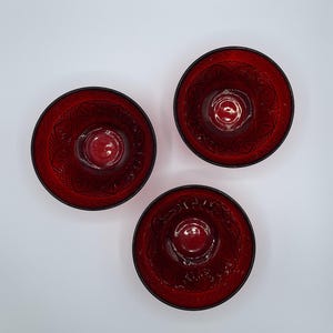 May include: Three ruby red glass bowls with black rims are arranged on a white surface. Each bowl has a detailed, etched floral pattern on the inside. The bowls are circular and appear to be empty.
