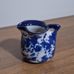 May include: A small, ceramic creamer with a blue and white speckled pattern. The creamer has a handle and a spout, and it sits on a wooden surface. The creamer is approximately 3 inches tall.