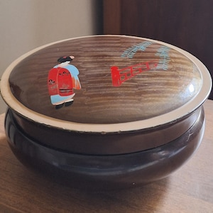 May include: A dark brown, oval-shaped trinket box with a cream-colored rim. The lid features a painted scene of a woman in a kimono and a red gate with a tree branch. The box is likely made of wood and has a glossy finish.