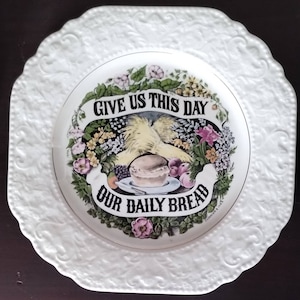 May include: White ceramic plate with a raised floral design border. The center of the plate features a colorful illustration of wheat, flowers, and a loaf of bread. The text "Give us this day our daily bread" is written in a banner around the illustration.