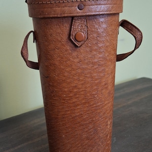 May include: A cylindrical brown leather case with a lid and two small handles. The case has a textured surface and a button closure on the front. The case is likely designed to hold a telescope or other similar item.
