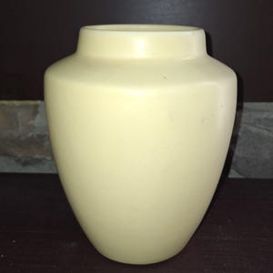 May include: A cream-colored vase with a rounded body and a slightly flared rim. The vase has a smooth, matte finish and a simple, elegant design. This decorative vase is suitable for displaying flowers or as a standalone accent piece.