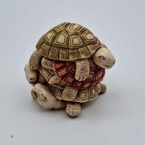 May include: A stacked turtle figurine with a beige and reddish-brown color scheme. The figurine has a detailed, textured shell design. The turtles are arranged in a playful, layered composition, creating a unique decorative piece.