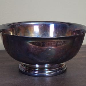 May include: A silver-colored metal bowl with a wide, rounded shape. The bowl sits on a circular base and has a reflective surface with hints of purple and blue. The bowl is likely used for serving or display.