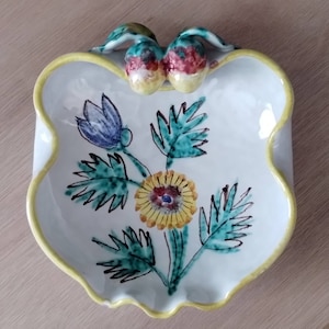 May include: A white ceramic dish with a scalloped edge and a yellow rim. The dish features a hand-painted floral design with green leaves, a yellow flower with a red center, and a blue flower. The dish has two small, yellow, ceramic knobs on the top.
