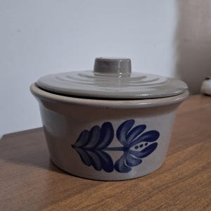 May include: A small, round, stoneware lidded container. The container is light gray with a blue floral design on the side. The lid is also light gray and fits snugly on top. The container is sitting on a wooden surface.
