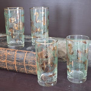 May include: Four vintage highball glasses. The clear glasses have a gold rim and are decorated with a gold and seafoam green pattern. The glasses are cylindrical and sit on a dark surface.