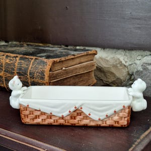 May include: A rectangular ceramic dish with a woven basket design, white interior, and a white cloth draped over the top, adorned with red cherry accents. Two white cat figurines serve as handles.