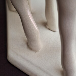 Pode incluir: Close-up de uma escultura de cer&acirc;mica creme com as pernas de uma figura. A escultura tem um acabamento craquelado e &eacute; montada em uma base retangular. As pernas s&atilde;o lisas e arredondadas.