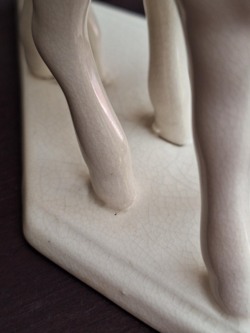 Pode incluir: Close-up de uma escultura de cer&acirc;mica creme com as pernas de uma figura. A escultura tem um acabamento craquelado e &eacute; montada em uma base retangular. As pernas s&atilde;o lisas e arredondadas.