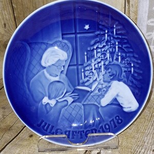 May include: A cobalt blue decorative plate featuring a Christmas scene. The plate depicts a woman reading to two children, with a Christmas tree and candles in the background. The text "Jule Aften 1978" is at the bottom.