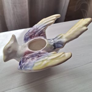 May include: A ceramic bird-shaped figurine featuring a glossy finish with blended pastel colors including soft yellow, light purple, and white. The bird is depicted in a flying pose with wings extended and a circular opening on its back.