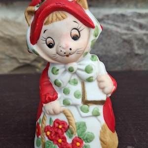 May include: A ceramic figurine of a cat-like character wearing a red hood and a white dress with green polka dots. The figurine holds a basket of red flowers and has a smiling face. The overall design is whimsical and decorative.