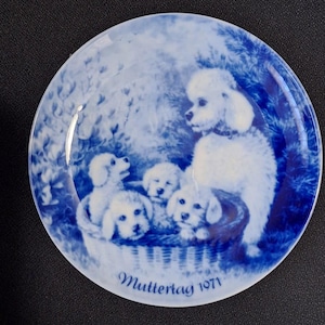 May include: A round, blue and white decorative plate featuring a poodle with four puppies in a basket. The plate has a floral background and the words "Muttertag 1971" are written at the bottom.
