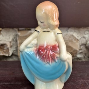 May include: A ceramic figurine of a girl in a blue dress, holding the skirt. The figurine has a peach-colored head and a red bow on the dress. The dress is a light blue color, and the figurine is on a dark brown surface.