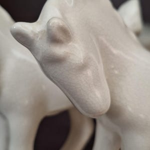 Pode incluir: Close-up de uma estatueta de cavalo de cer&acirc;mica creme. A cabe&ccedil;a do cavalo est&aacute; inclinada para baixo, com detalhes vis&iacute;veis da orelha e do focinho. A cer&acirc;mica tem um acabamento craquelado, adicionando textura &agrave; escultura. O fundo &eacute; de cor escura e neutra.