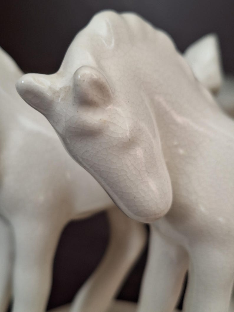 Pode incluir: Close-up de uma estatueta de cavalo de cer&acirc;mica creme. A cabe&ccedil;a do cavalo est&aacute; inclinada para baixo, com detalhes vis&iacute;veis da orelha e do focinho. A cer&acirc;mica tem um acabamento craquelado, adicionando textura &agrave; escultura. O fundo &eacute; de cor escura e neutra.