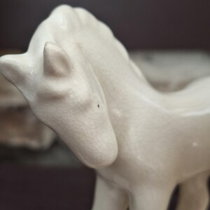 Pode incluir: Estatueta de cavalo em cer&acirc;mica na cor creme. O cavalo est&aacute; em p&eacute; com a cabe&ccedil;a ligeiramente virada para o lado. A estatueta tem uma superf&iacute;cie texturizada e &eacute; um objeto decorativo.