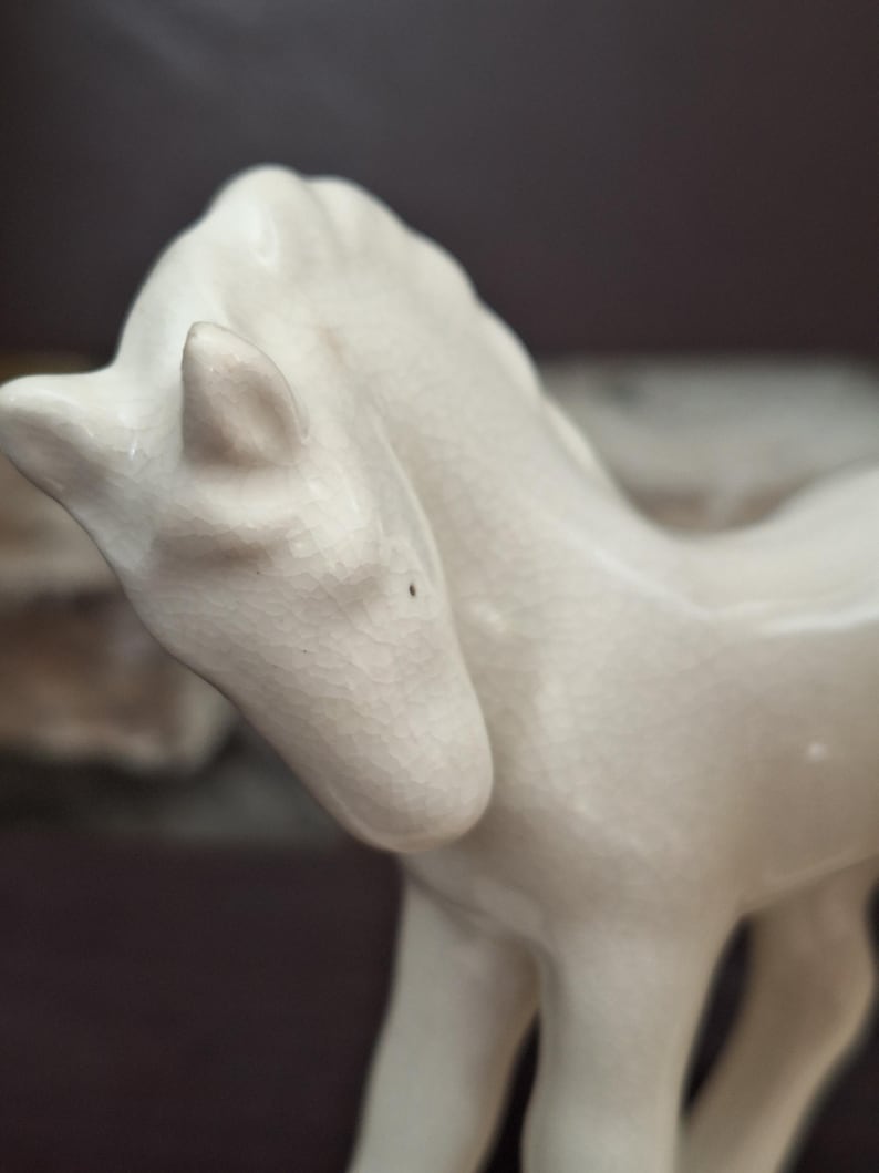 Pode incluir: Estatueta de cavalo em cer&acirc;mica na cor creme. O cavalo est&aacute; em p&eacute; com a cabe&ccedil;a ligeiramente virada para o lado. A estatueta tem uma superf&iacute;cie texturizada e &eacute; um objeto decorativo.