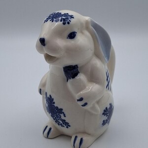 May include: A ceramic creamer shaped like a rabbit with a glossy white finish and blue floral patterns. The rabbit features blue painted details on its ears, eyes, and paws, and it holds a small carrot. The piece has a spout at the mouth area.