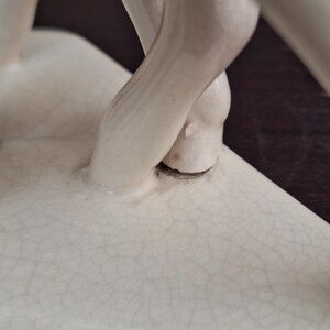 Pode incluir: Close-up de uma escultura de cer&acirc;mica creme com rachaduras vis&iacute;veis. A escultura apresenta uma figura com as pernas cruzadas nos tornozelos. As pernas s&atilde;o lisas e a escultura tem um acabamento fosco. O fundo &eacute; marrom escuro.