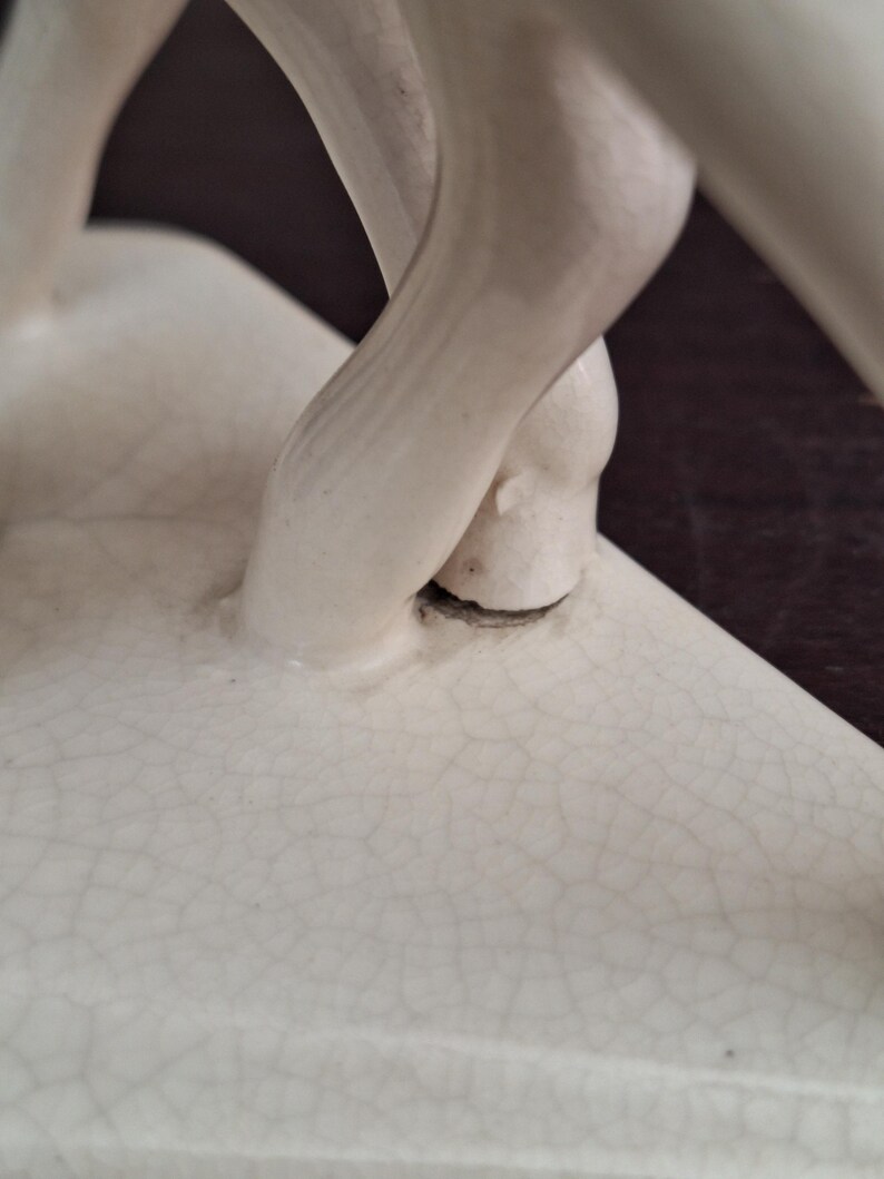 Pode incluir: Close-up de uma escultura de cer&acirc;mica creme com rachaduras vis&iacute;veis. A escultura apresenta uma figura com as pernas cruzadas nos tornozelos. As pernas s&atilde;o lisas e a escultura tem um acabamento fosco. O fundo &eacute; marrom escuro.