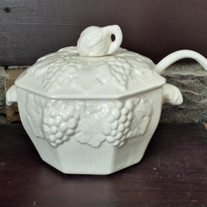 May include: Cream-colored ceramic soup tureen with a lid and ladle. The tureen is octagonal and decorated with grape clusters. The lid has a handle shaped like a grape leaf. The ladle rests on the side of the tureen.
