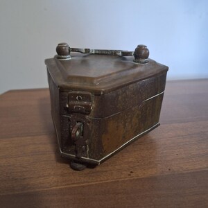 May include: A small, antique bronze-colored box with a triangular shape. The box has a hinged lid, a handle, and a latch with a keyhole. The box is sitting on a wooden surface.