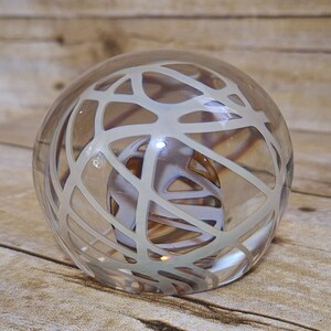 May include: A clear glass paperweight with a white web-like design inside. The paperweight is round and has a smooth surface.