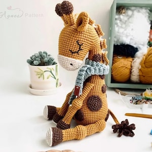 May include: A crocheted giraffe toy with brown spots and a blue scarf. The giraffe is sitting on a white surface with a small potted plant and yarn in the background.
