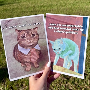 May include: Two rectangular greeting cards featuring cat illustrations. The left card shows a brown cat wearing a white collar and red striped tie with the text i don't want to work. The right card shows a light blue cat with the text When i'm uncomfortably hot and someone asks me a stupid question.