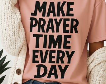 May include: A light pink t-shirt with the text "MAKE PRAYER TIME EVERY DAY" in bold black letters. The person is wearing blue jeans and a cream-colored cardigan.