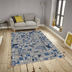 May include: A rectangular rug with a detailed geometric pattern in blue, white, and grey. The design resembles a technical blueprint or a futuristic city map. The rug is placed on a wooden floor in a living room setting, with a sofa and window visible.