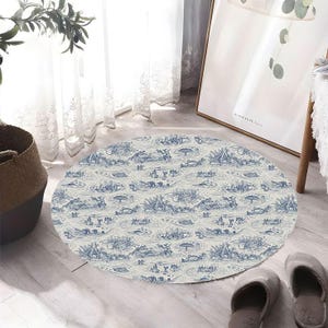 Puede incluir: Una alfombra redonda con un diseño de toile de Jouy azul y blanco. El patrón presenta una escena pastoral con figuras, árboles y elementos acuáticos. La alfombra está sobre un suelo de madera clara, con zapatillas cerca.