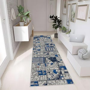 May include: A rectangular hallway rug with a geometric design in shades of blue, grey, and white. The rug features a detailed pattern resembling a spaceship, with a circular design at one end. The rug is placed on a white floor.