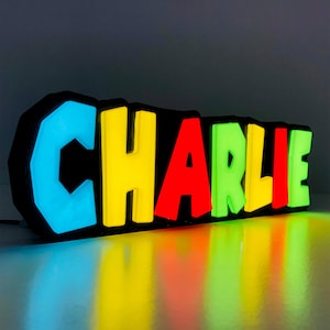 May include: A colourful, illuminated name sign spelling "CHARLIE" with each letter in a different colour: blue, yellow, red, green, yellow, and red. The sign is made of plastic and has a black background.