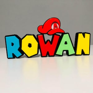 May include: A vibrant 3D-printed name sign spelling "ROWAN" in block letters. The letters are outlined in black with bright colours: blue, yellow, red, green, and yellow. A red hat with a white "M" sits above the "W".