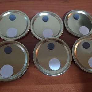 May include: Six circular gold-colored metal lids arranged in two rows. Each lid features a small, centered blue circular accent near the top edge and a larger, solid white circular sticker positioned toward the bottom edge of the metallic surface.