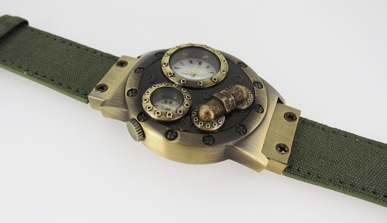 K&ouml;nnte beinhalten: Eine goldfarbene Uhr mit einem mehrschichtigen Zifferblatt und einem gr&uuml;nen Canvas-Armband. Das Zifferblatt der Uhr hat drei runde Zifferbl&auml;tter, eines mit r&ouml;mischen Ziffern und zwei mit arabischen Ziffern. Die Uhr hat einen Steampunk-Look.