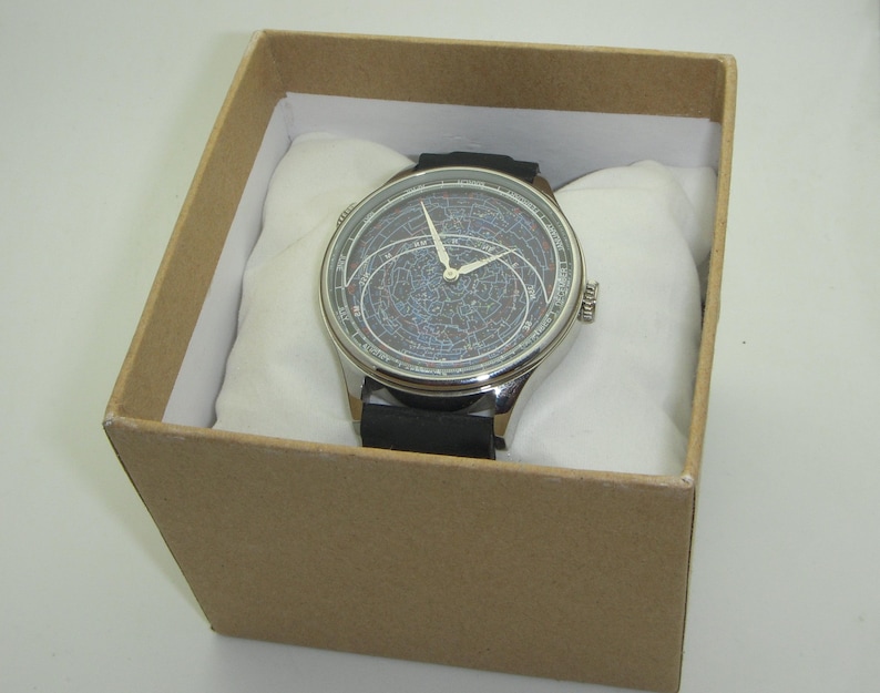 May include: A silver watch with a black leather band and a unique, detailed, circular face. The watch is in a brown box with white padding.